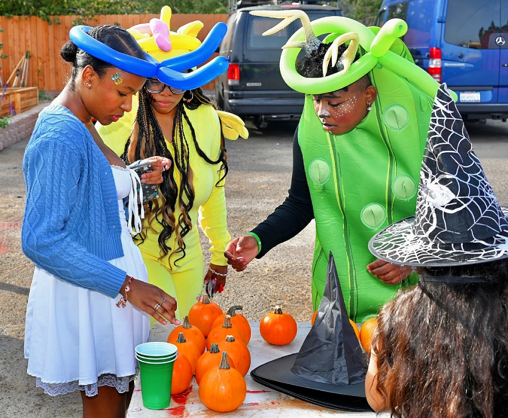 Three volunteers in costumes line up pumpkins in front of a witch hat.