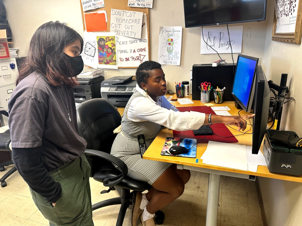 One woman points to a computer screen teaching another woman