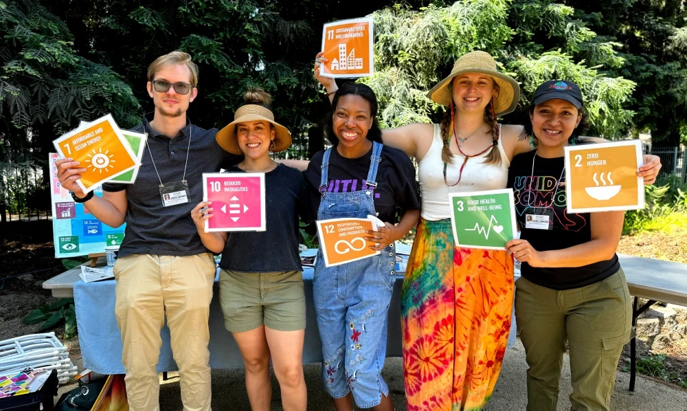 5 people stand together smiling and holding posters with Sustainable Development Goal Icons.