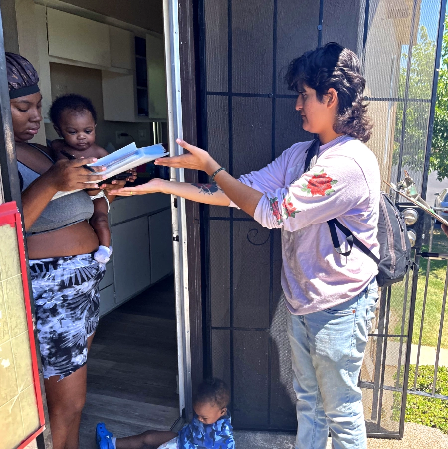 Woman stands at door with baby. Volunteer hands woman clipboard with papers.