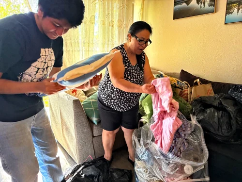 Man takes pillow out of a bag. Woman next to him takes clothing out of a bag.