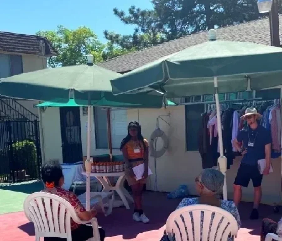 Group of people sitting in a patio. Volunteers standing with clipboards presenting. Clothing rack set up in the background.
